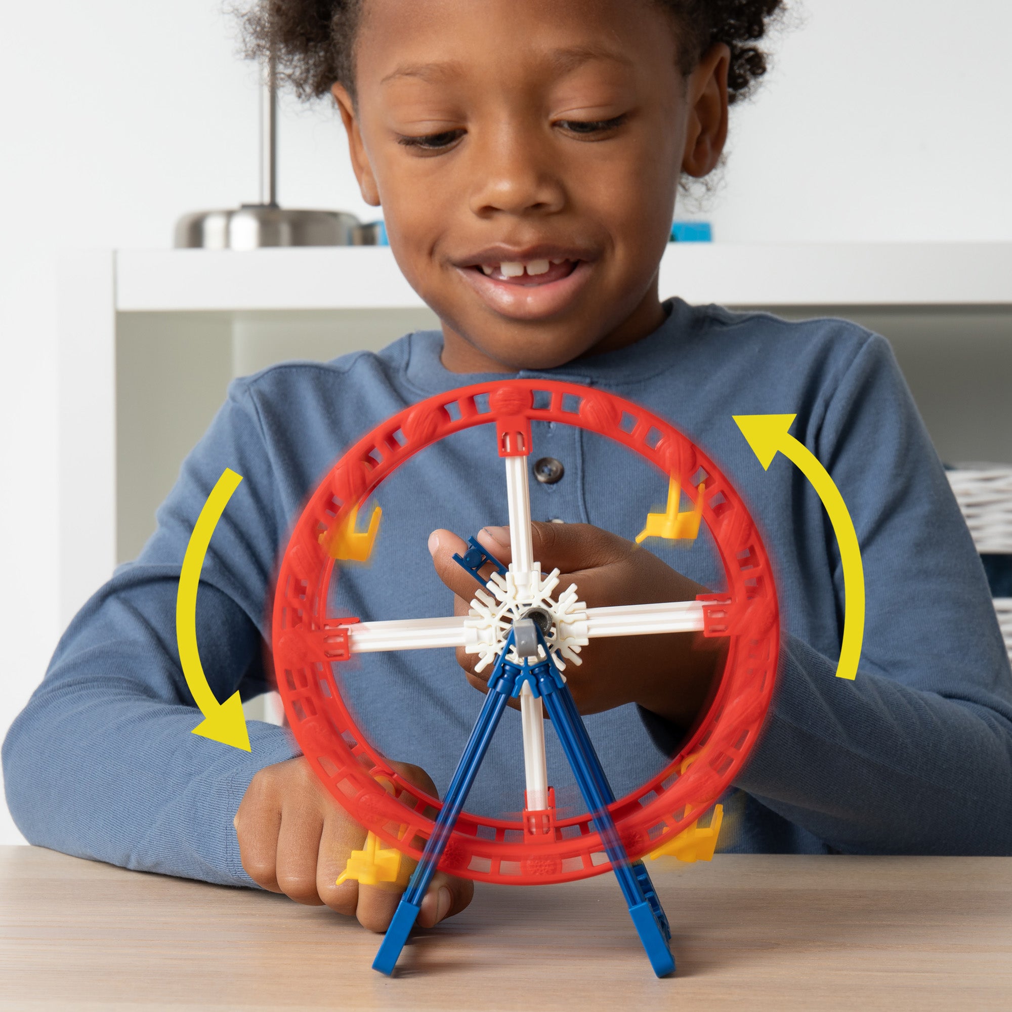 A smiling child wearing a blue long-sleeve shirt is playing with a small model Ferris wheel. The Ferris wheel features red, blue, and white components and is positioned on a table. Yellow arrows are shown indicating the rotation direction of the Ferris wheel.