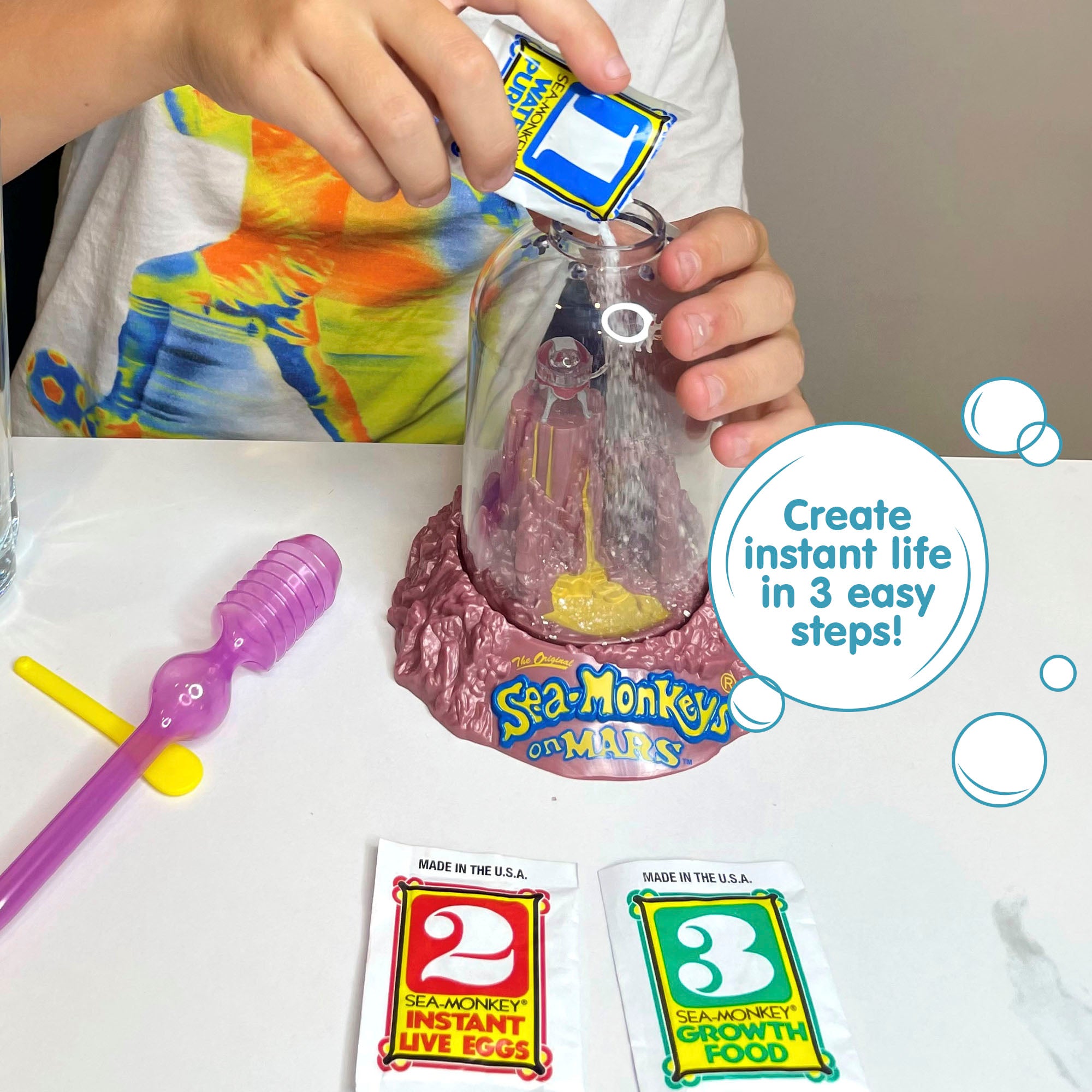 A child wearing a white shirt with bright colors printed on it is holding the Magiquarium with one hand, and pouring powder from a small packet labled with the number 1 with the other hand. A packet with the number 2, a packet with the number 3, a purple pipette, and a yellow plastic spoon all lay on the surface in front of the child. There is a callout in a circle that reads "Create instant life in 3 easy steps!".